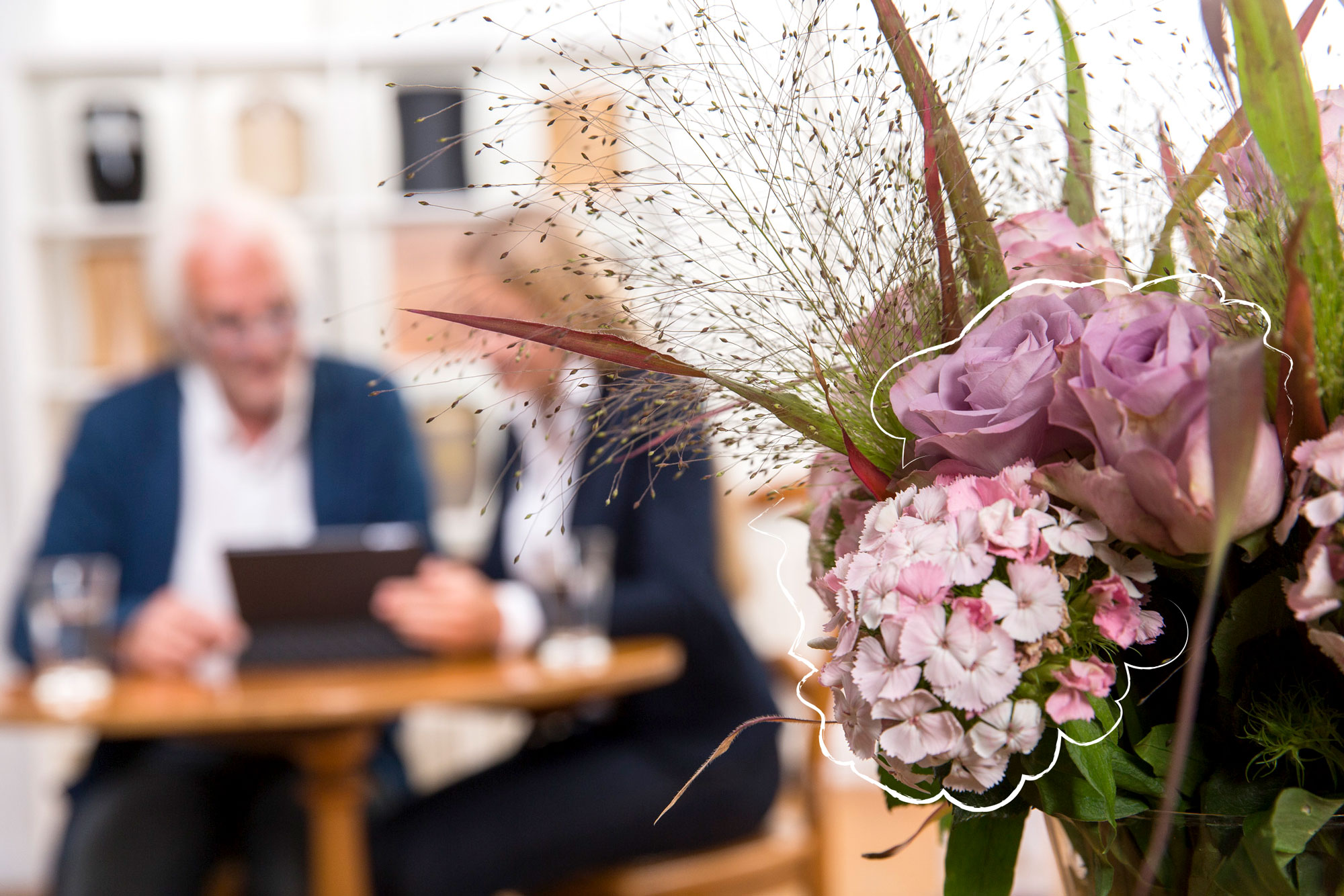 Beratungsgespräch an einem Tisch, im Vordergrund ein Blumenstrauß mit Rosen und Nelken